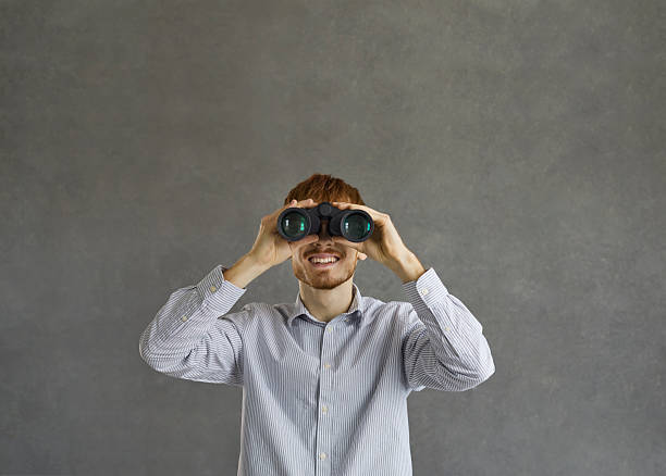 Portrait of happy young man holding optical zoom binoculars and looking ahead standing against grey studio background. Searching for job, making discoveries, looking forward to something concept