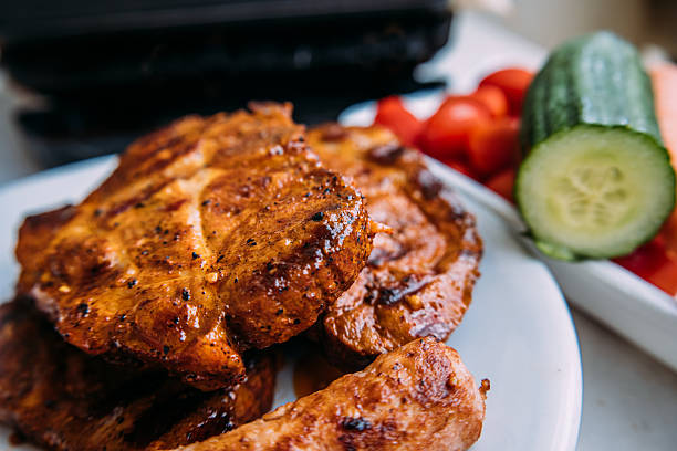 Juicy grilled pork steaks on a plate with sliced cucumbers and tomatoes, perfect for summer barbecue serving.