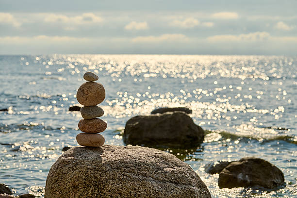 Stack of smooth stones precariously balanced on larger rock by calm seaside, symbolizes balance and serenity, evoking peaceful and meditative atmosphere