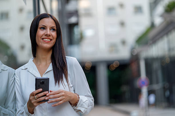 istock Young businesswoman smiling while engaging with her smartphone, surrounded by sleek modern office buildings in an urban setting 2229774463