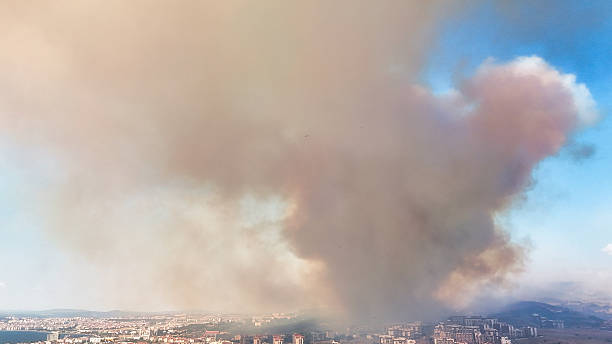 Drone photograph capturing a massive smoke column rising above urban buildings due to a nearby fire. The dense, multi-colored smoke contrasts with the blue sky, highlighting the impact of large-scale fires on city environments and air quality.
