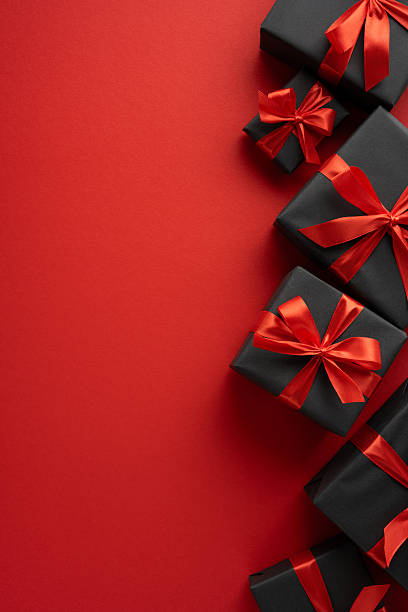 istock Beautifully wrapped black gift boxes adorned with red ribbons, placed on a vertical bold red backdrop, conveying elegance, festive celebration, and style 2229790871