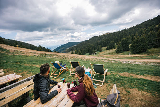 Two hikers relax in a mountaintop restaurant after a hike, drinking fresh juice and enjoying a stunning panoramic view of nature.