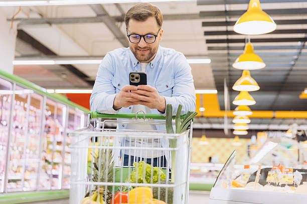 istock Smiling customer using smartphone while shopping in supermarket with full cart 2229793818