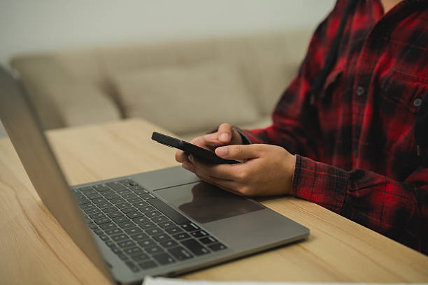 istock Person in Red Flannel Shirt Using Smartphone While Working on Laptop at Home Office with Cozy Background 2229836410