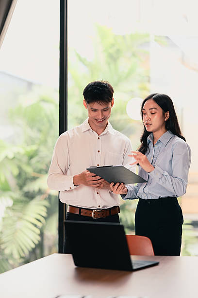 istock Two young Asian colleagues standing by a large window, discussing work while reviewing documents 2229838838