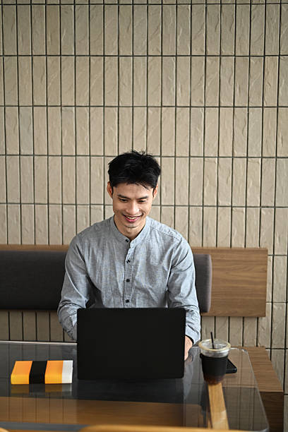 istock Young Asian man working on a laptop at a modern cafe table with a drink and notebook 2229838856