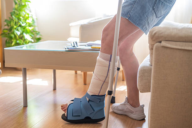 istock Mature woman is standing up from a sofa in her living room, using crutches for support while wearing an orthopedic boot on her injured leg, recovering from a metatarsal fracture 2229852657