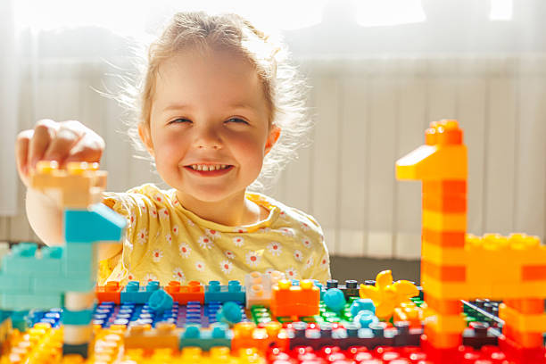 istock A beaming toddler girl with a messy bun smiles brightly at the camera while playing with colorful building blocks. 2229866111