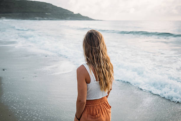 Young woman with long hair standing on sandy beach facing ocean waves. Casual summer outfit by the sea. Person enjoying coastal scenery and peaceful waterfront view.