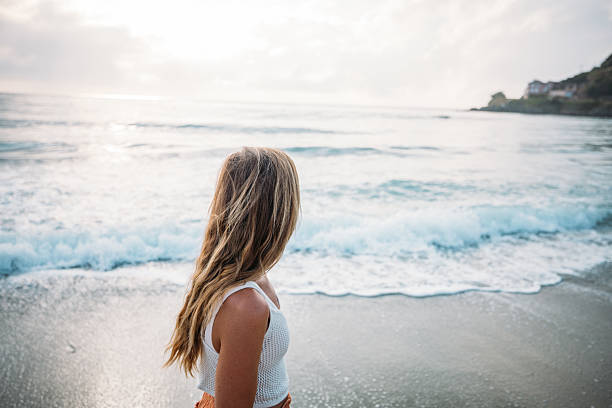 Young woman with long hair standing on sandy beach facing ocean waves. Casual summer outfit by the sea. Person enjoying coastal scenery and peaceful waterfront view.