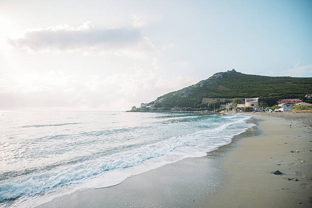 Gentle waves washing onto sandy beach near green hills and small buildings. Peaceful coastal landscape with soft sunlight and cloudy sky. Scenic ocean shore with natural and urban elements.