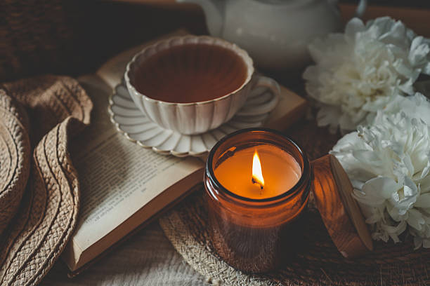 istock Lit candle on a rustic table with a cup of tea, white peonies, an open book, and a straw hat. Warm, cozy flat lay with vintage tones and soft natural light 2229880906