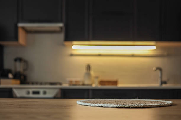 istock Cozy modern kitchen interior with a round textured placemat on a wooden table in soft focus foreground. Minimalist design with dark cabinets and under-cabinet lighting in the background 2229881011