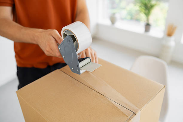 istock Person sealing a cardboard box with tape in a bright indoor setting 2229882413