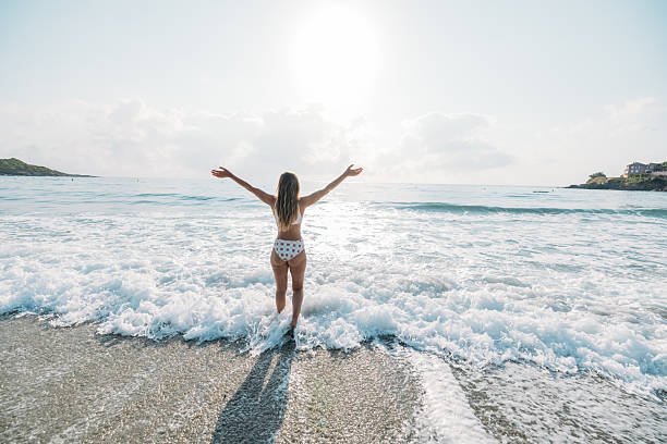Young woman in polka dot swimsuit walking into foamy ocean waves with arms raised. Carefree beach day with bright sunlight and gentle sea breeze. Relaxing summer vacation by the water.
