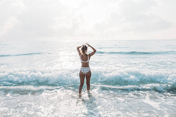 Woman in polka dot swimsuit standing at the edge of the ocean with waves crashing. Bright sunny day on a sandy beach with clear water. Beach vacation and summer relaxation concept.
