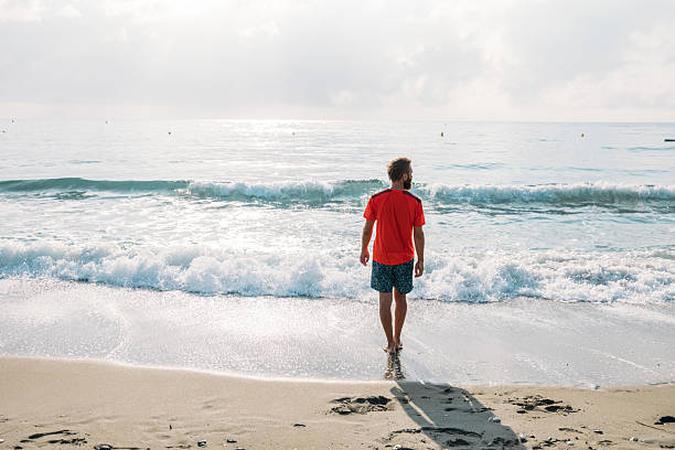 Bearded man in orange shirt and shorts walking barefoot along the sandy beach near gentle ocean waves. Casual summer beach scene with peaceful coastal landscape and green hills in background. Outdoor seaside relaxation.