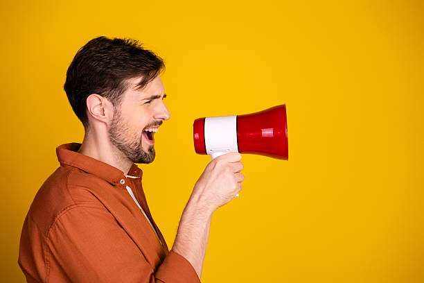 istock Young man speaking through red megaphone against vibrant yellow background, showcasing advertising and communication concepts 2229896409
