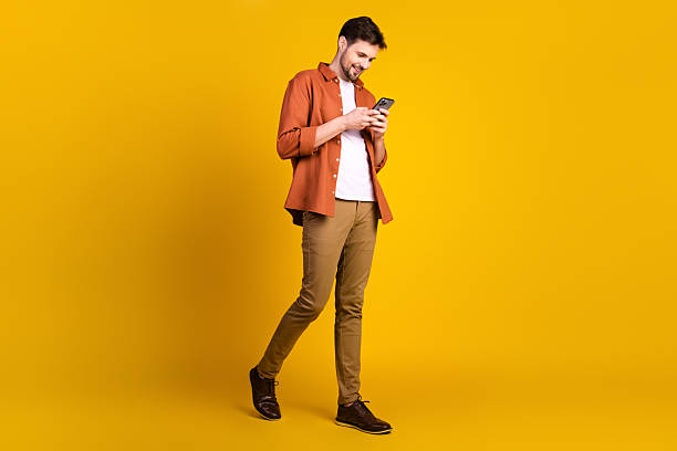 istock Modern young man in casual outfit using smartphone while standing near vibrant yellow background indoors 2229896417