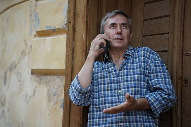 A mature man standing by an old wall, using his smartphone outdoors. He appears relaxed and thoughtful, enjoying a peaceful moment. Ideal for themes like digital lifestyle, communication, and everyday life.