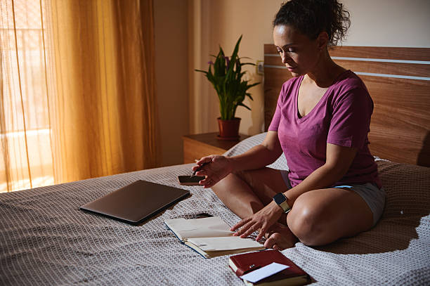 istock Woman Sitting on Bed Planning Her Day Using Notebook and Smartphone 2229902366