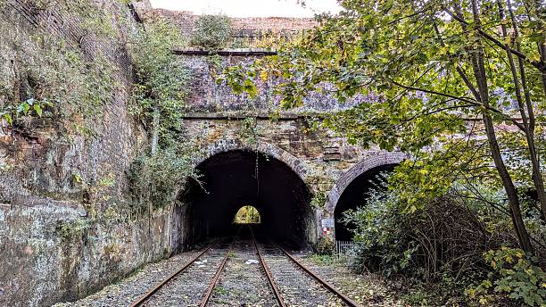 istock Derelict twin dark stone railway tunnels with rail tracks underneath large brickwork bridge with cliff walls, trees, and overgrown bushes of abandoned train line 2229913447
