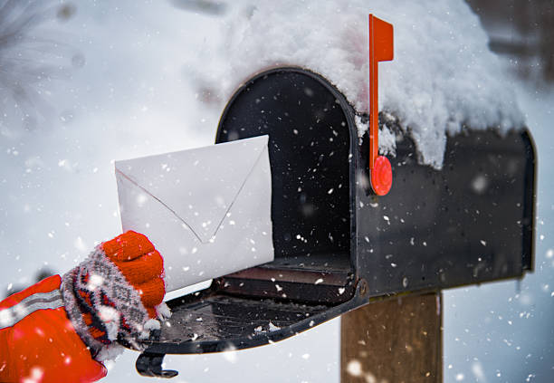 Close-up of a gloved hand placing a white envelope into a black mailbox topped with snow. Snowflakes fall around, creating a cozy winter atmosphere, with the bright red flag of the mailbox standing out against the snowy background.