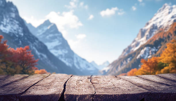 Rustic wooden table in sharp focus with a blurred background of majestic snow-capped mountains and colorful autumn trees. The scenic alpine landscape evokes a peaceful, seasonal atmosphere, perfect for product placement, travel, nature, or food-related designs.