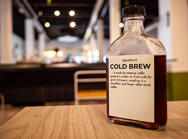 Close-up of a clear glass bottle labeled “Signature Cold Brew,” placed on a wooden table in a softly blurred cafe setting. The label highlights the 16-hour steeping process that creates a smooth flavor with a lasting aftertaste.