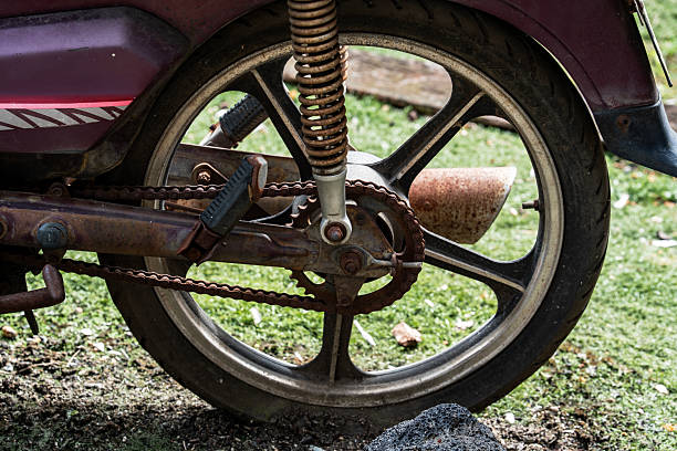 Close-up of a motorcycle’s rear wheel, chain, and exhaust pipe, showing heavy rust and wear, set against a backdrop of green grass and dirt.