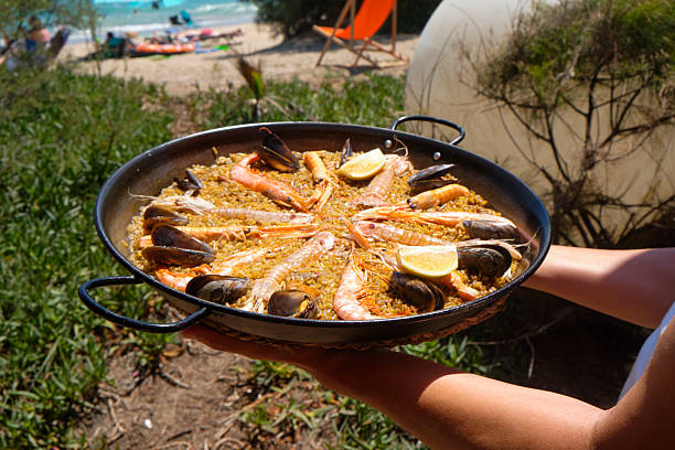 a seafood paella takes center frame in a frontal view with mussels and prawns glistening in the sun as a beach scene with umbrellas appears in soft focus behind