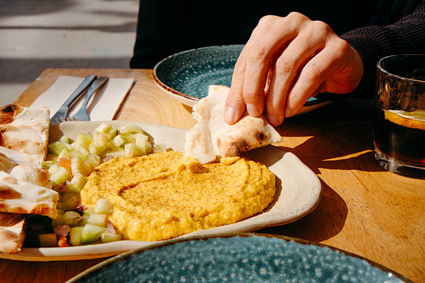 a man scoops creamy curry hummus with a piece of warm naan bread served alongside fresh diced salad on a wooden table at a sunny sidewalk cafe in the city