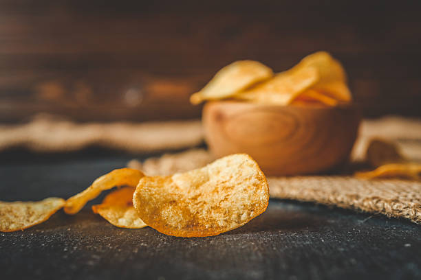 istock Crispy potato chips in a rustic wooden bowl on burlap fabric, with scattered chips on a dark textured table. Cozy snack scene with natural tones 2229936400