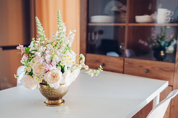 Fresh flower arrangement made in brass vase put on dining table decorating kitchen interior. Bouquet made of white beige roses, digitalis blooms left by cupboard with tableware