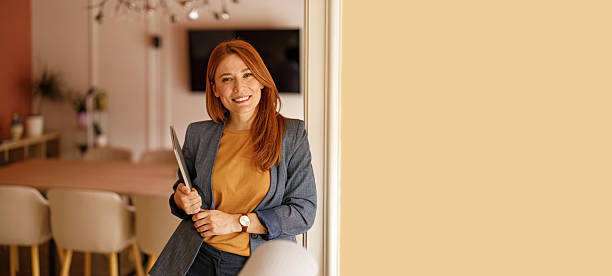 istock Smiling businesswoman holding computer and looking confidently at camera while leaning on wall at board room 2229943248