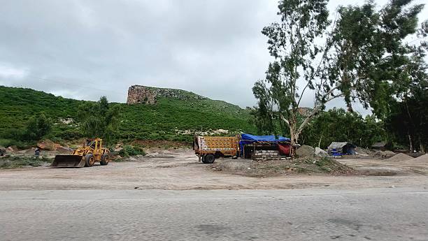 Construction vehicles rest in a rural area. A yellow front loader and a truck are parked on a dirt surface. Nearby, a blue and red tarped structure is visible. The scene is bordered by a grassy hill, partially covered with shrubs, and dotted with trees, including a prominent eucalyptus. Overhead, a cloudy sky stretches across the landscape, casting a muted light. The setting suggests industrial activity amidst a natural, verdant backdrop.