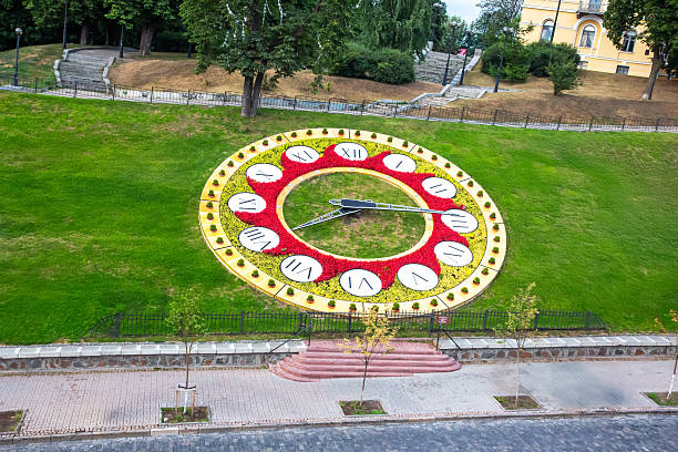 A vibrant clock installation made of flowers and greenery sits prominently on a hillside in a city park, showcasing Roman numerals and large clock hands against a bright background.
