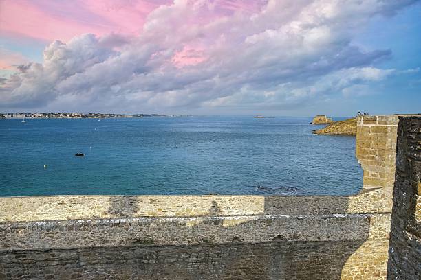 A scenic coastal view from stone fortifications overlooks calm blue water under a dramatic pink and purple sunset sky.