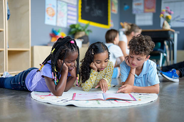 Three children are lying on a colorful rug in a classroom, engaged in reading and sharing a book together. The scene conveys collaboration, fun learning, and friendship in an educational environment.
