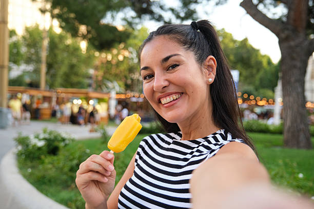 istock Young woman taking a selfie while eating popsicle in a park 2229954315
