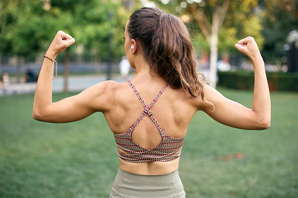 A physically fit woman displays her toned arms while flexing outdoors, wearing stylish activewear. The image captures strength, health, and determination in a sunny and green natural environment.