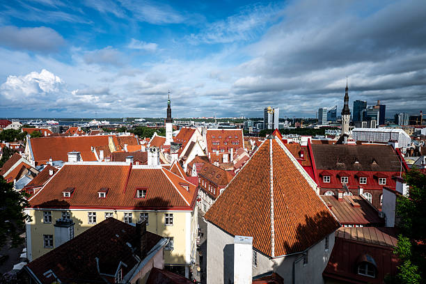Scenic panoramic view of Tallinn’s Old Town, Estonia, featuring red-tiled rooftops, historic spires, and the distant modern skyline, with the Baltic Sea and ferry terminals visible on the horizon under a partly cloudy sky.