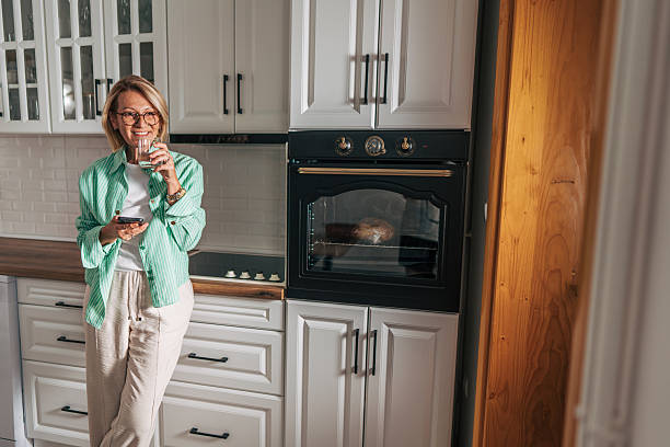 istock Smiling woman drinking water and using smartphone in kitchen 2229964382