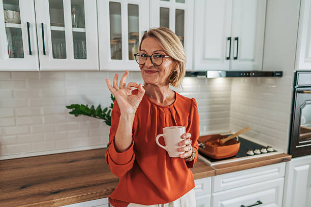 Smiling senior woman gesturing ok while drinking coffee in kitchen