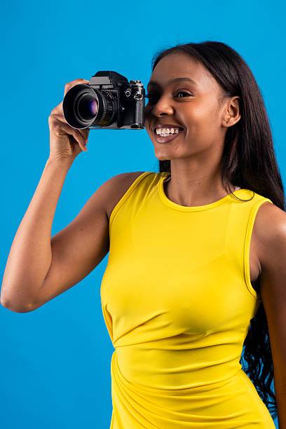 Young black woman photographer smiling and taking photos with a professional analog camera in studio over blue background