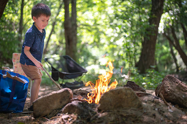 A young boy interacts closely with a campfire, showcasing his curiosity and adventurous spirit in a lush green camping setting while maintaining focus on the flames before him.
