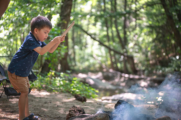 A young child is focused and determinedly blowing on the embers of a campfire with a piece of cardboard to help the flames grow. The scene captures a moment of childhood learning and participation in an outdoor activity, highlighting themes of fire-starting, camping, and youthful curiosity in a natural environment.