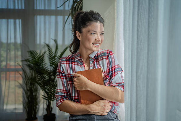 istock Young woman relaxing by window, holding a book and smiling 2229983653