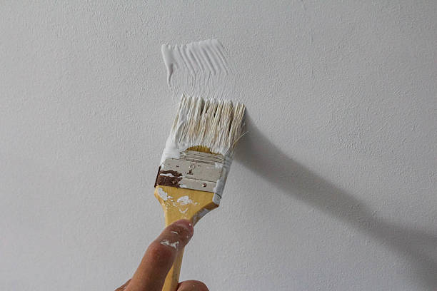 istock Paint brush with wooden handle applying white paint to wall surface during professional interior renovation project 2229994961